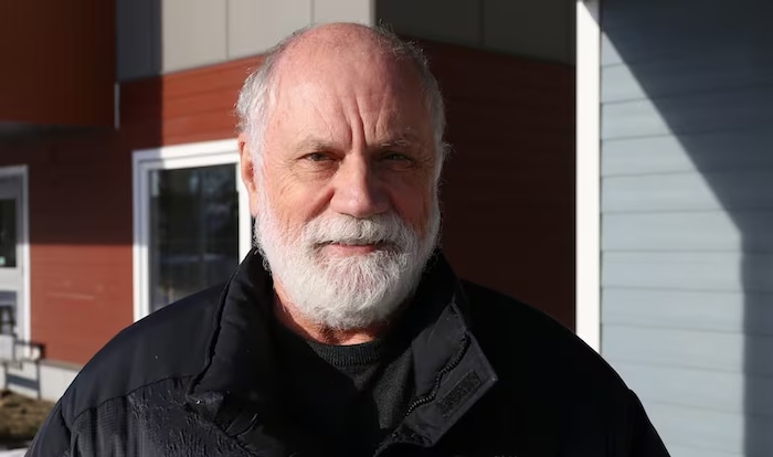 Une personne portant une barbe et une moustache blanches regarde dans l'objectif d'une caméra.