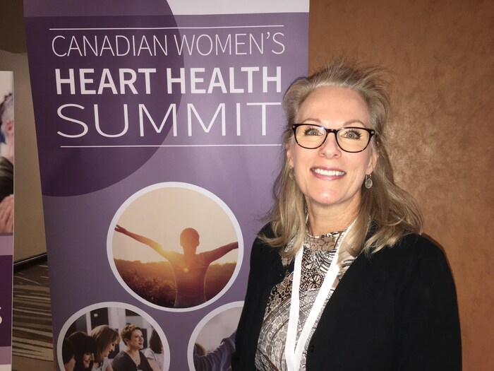 Une dame sourit à la caméra devant une bannière du Sommet canadien sur la santé cardiaque des femmes.