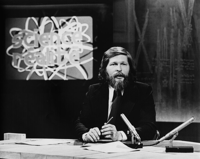 Dans un studio de télévision, l'animateur Joël Le Bigot est assis à une table
surmontée d'un micro. En arrière-plan, le titre de l'émission « Science Réalité » apparaît
dans un écran sur le mur.