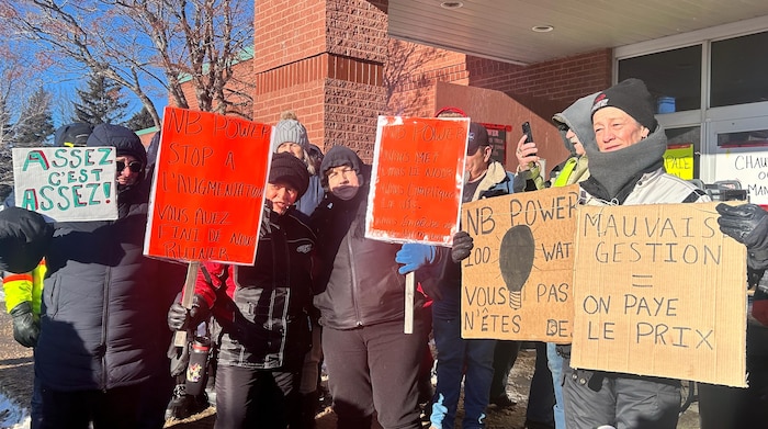 Des gens dehors en hiver posent avec des pancartes, avec des messages à la clarté discutable, comme «NB Power 100 watts vous pas n'êtes des».