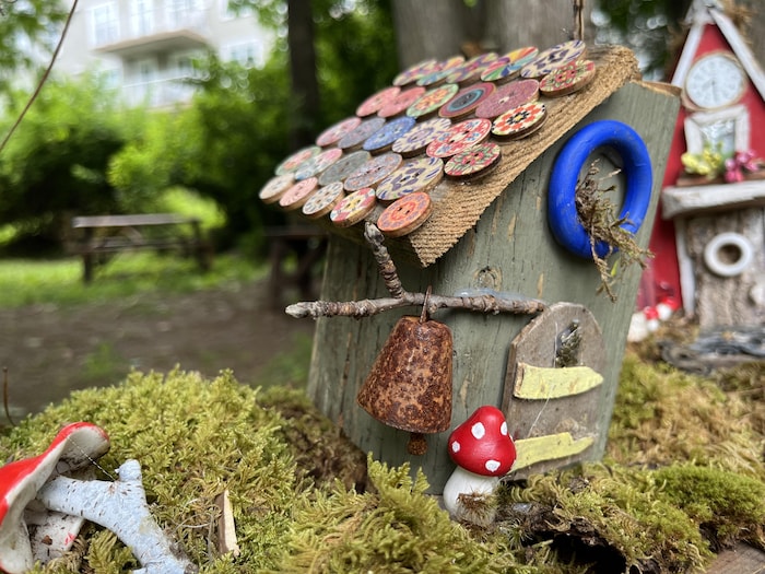 Une cabane miniature au toit en pente décoré de boutons, avec une cloche rouillée et un champignon rouge et blanc devant.