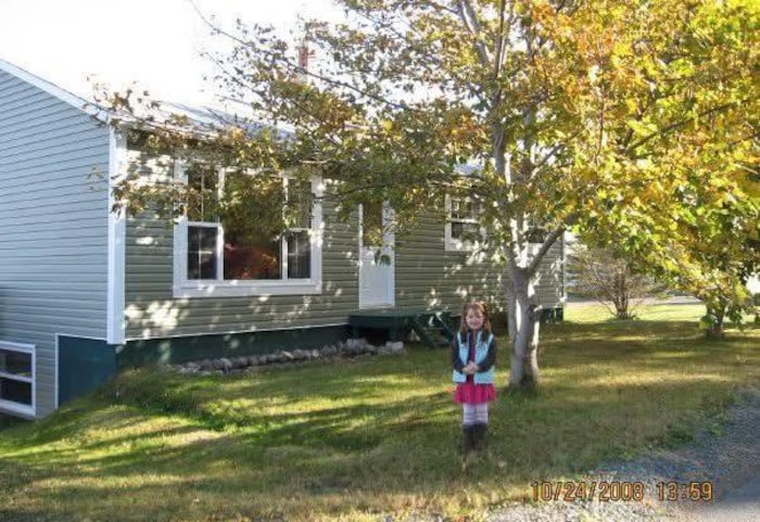 Photo d'une petite fille debout dans la cour devant sa maison et devant un arbre. La date du 24 mars 2008 est inscrite dans le coin inférieur droit.