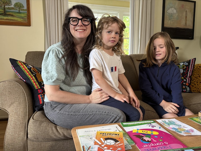Une jeune mère portant des lunettes à grosses montures est assise sur un sofa avec un enfant sur les genoux, et un autre assis à côté.