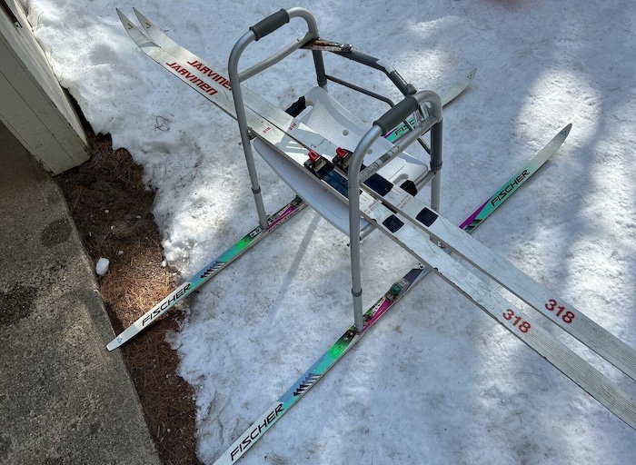 
Une marchette avec des skis posés en dessous, de chaque côté. La marchette a aussi un siège pour s'assoir. Deux autres skis de fond sont posés sur le banc.