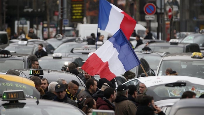 Des manifestants tiennent des drapeaux français entre plusieurs voitures de taxi à l'arrêt.