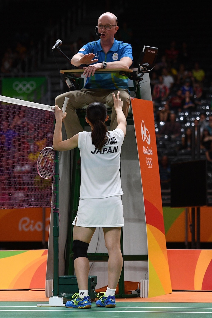 A referee sitting on a high chair, shows the palm of her hand to a player, standing and back to the camera, who makes the sign of the number 'two' with her fingers.
