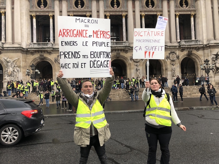 Un homme et une femme portant des gilets jaunes brandissent des pancartes dénonçant l'arrogance d'Emmanuel Macron.