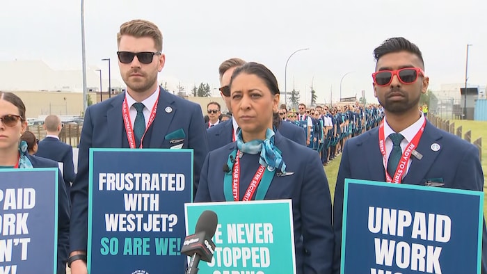 Trois agents de bord de la compagnie WestJet en uniforme, portant des lunettes de soleil et des cordons rouges, tiennent des pancartes de manifestation syndicale lors d'un point de presse.