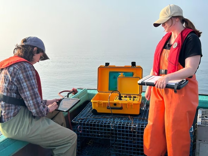 Deux femmes sur un bateau de pêche. Une batterie orange est posée sur un casier. Une des deux techniciennes utilise une tablette pour envoyer des signaux.