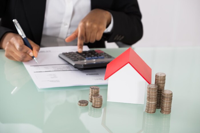 Un homme effectue des calculs et devant lui se tient une petite maison avec des pièces de monnaie.