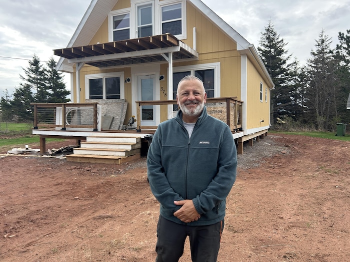 Un homme debout et souriant devant un chalet aux murs jaunes.