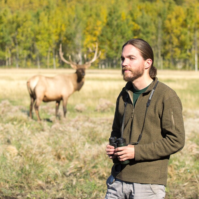 Jake Paleczny, des jumelles à la main, devant un caribou qui est en flou, en arrière plan.