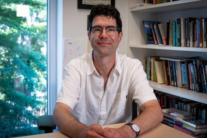 A man sits in an office with shelves of books and a window in backdrop.