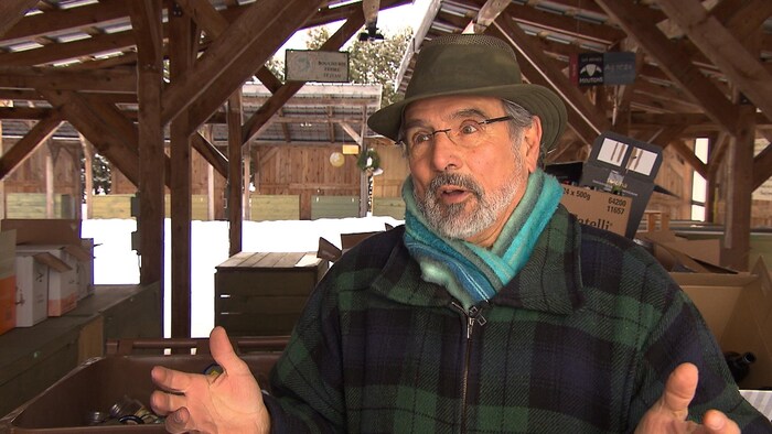 Un homme portant un chapeau, un manteau et un foulard gesticule avec ses bras en parlant avec un journaliste lors d'une entrevue à l'extérieur.
