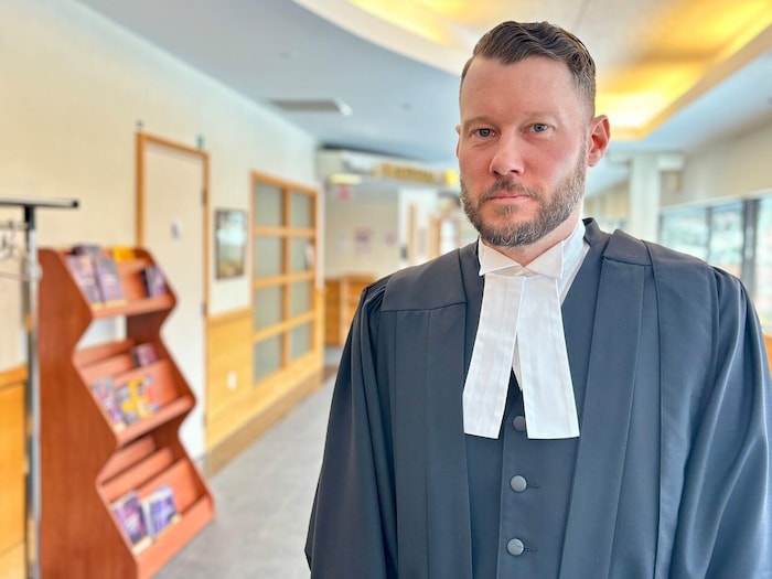 Un avocat portant la toge, debout dans un couloir du palais de justice. Il regarde la caméra.