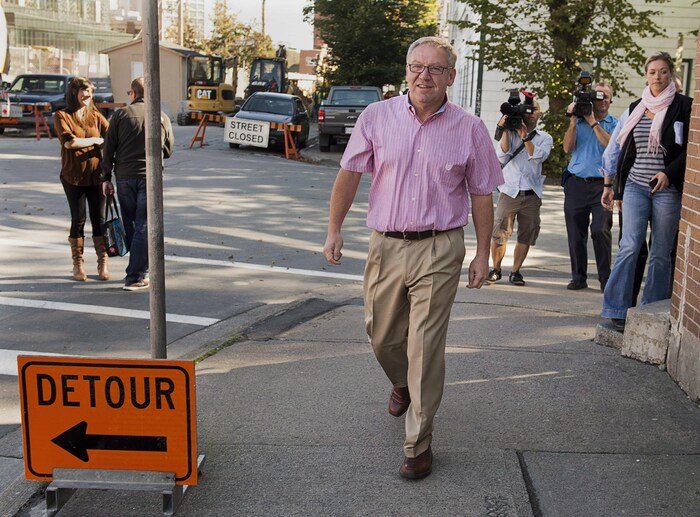 Suivi par des personnes qui marchent en le filmant avec de grosses caméras, un politicien en chemise rose à manches courtes marche sur le trottoir d'une rue fermée à la circulation.