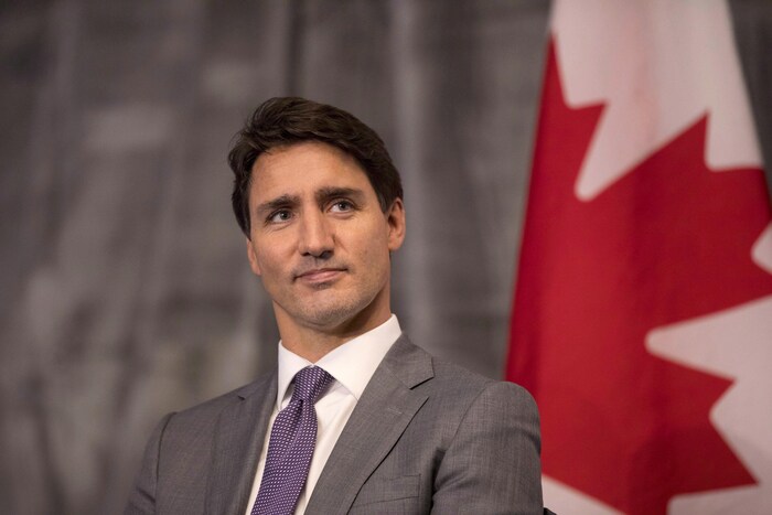 Le premier ministre Justin Trudeau regarde au loin. Derrière lui est accroché un drapeau du Canada. 
