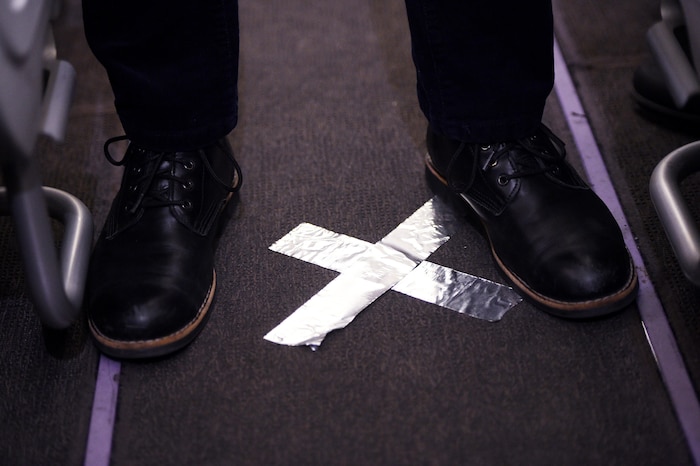 Deux chaussures foncées d'homme placées autour de deux morceaux de papier collant argenté formant un X sur le tapis de l'allée centrale d'un avion.