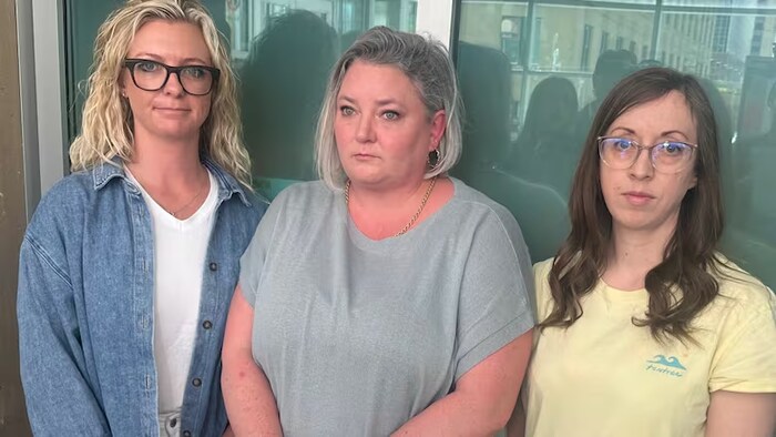 Trois femmes se tiennent debout, épaule contre épaule, en fixant du regard l'objectif d'une caméra.