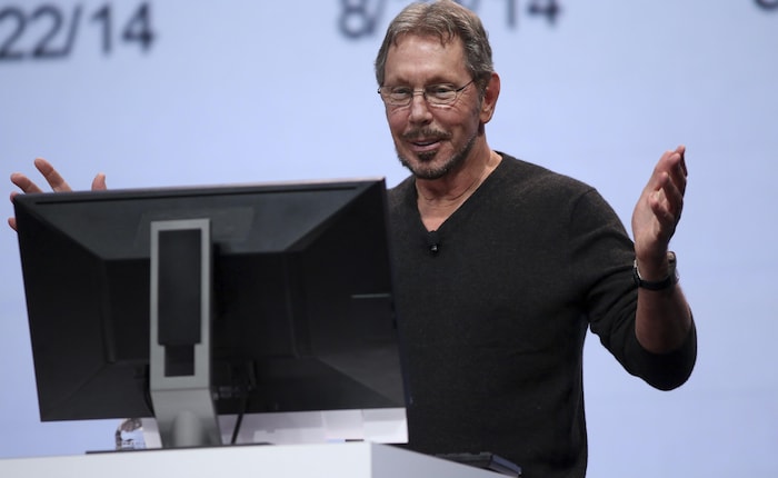 Larry Ellison étend les bras. Il est sur une scène face au moniteur d'un ordinateur, avec un grand écran derrière.