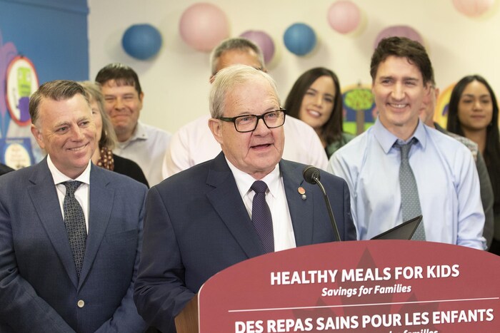 Un groupe de personnes dans une école dont le mur est décoré de ballons roses et bleus. Lawrence MacAulay est au micro devant un lutrin et parle, tandis que Dennis King et Justin Trudeau, de chaque côté de lui, sourient.