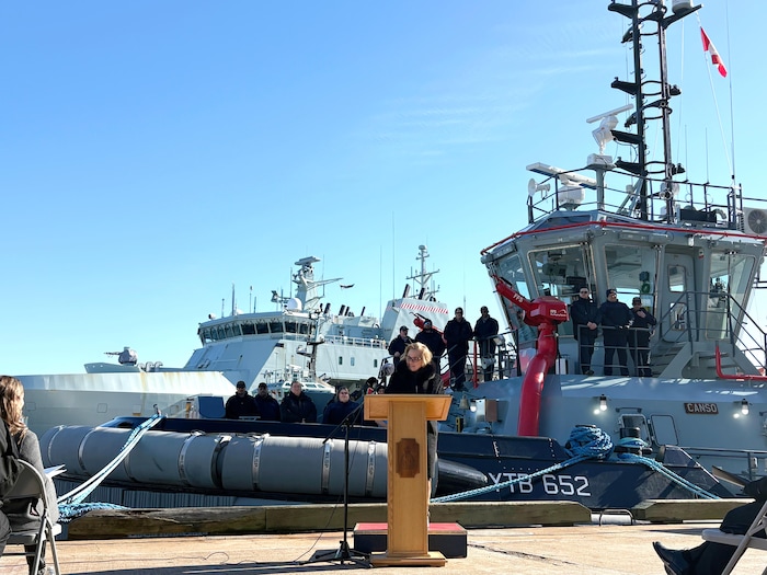Une femme sur le quai parle debout devant un lutrin. Elle est devant un remorqueur naval sur lequel prennent place plusieurs marins.