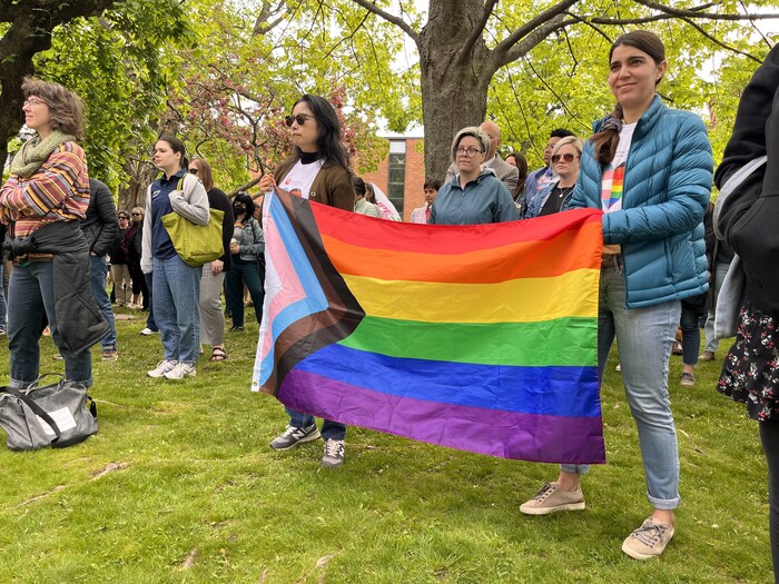 Des gens sont rassemblés sur le gazon. Deux personnes tiennent un drapeau arc-en-ciel.