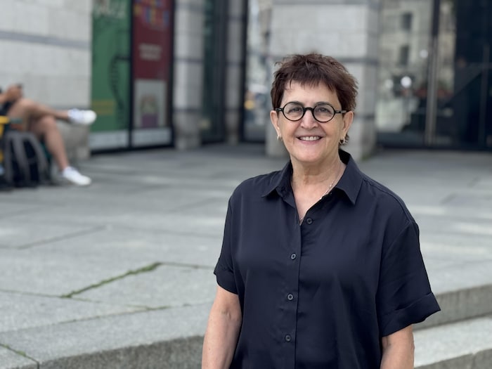 Une personne souriante, vêtue d’une chemise sombre et portant des lunettes rondes, pose devant un édifice moderne.