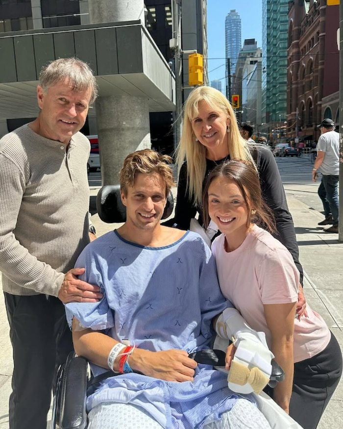 Lucas Arsenault, en fauteuil roulant et habillé de la chemise d'hôpital, entouré de sa famille souriante. 