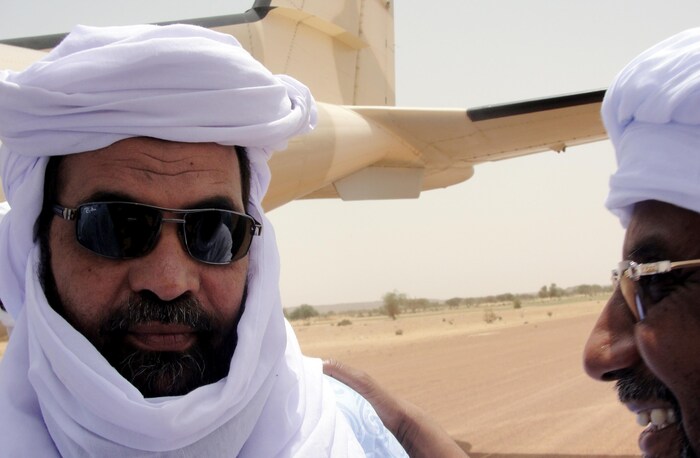 Un homme portant des lunettes de soleil, la barbe et un chèche blanc discute avec un autre homme vêtu de la même façon près d'un avion, au Mali.