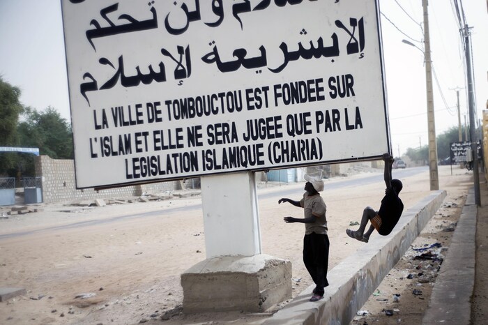 Deux jeunes Maliens se balancent sous une affiche blanche sur laquelle il est écrit en arabe et en français : « La ville de Tombouctou est fondée sur l'Islam et elle ne sera jugée que par la législation islamique (charia) ».