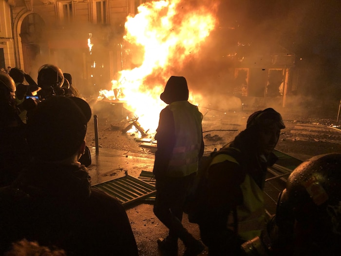 Des manifestants portant des gilets jaunes observent une barricade en feu dans une rue de Paris pendant la nuit.