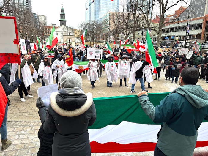 Neuf personnes, les vêtements couverts par des draps blancs tachés de rouge pour symboliser le sang, sont sur une place publique, entourés d'un grand nombre de manifestants. Plusieurs personnes tendent des drapeaux de l'Iran.