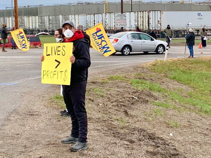 Un manifestant porte un masque sur le visage et une pancarte dans la main. On peut y lire en anglais que les vies valent plus que les profits. Il y a d'autres personnes à l'entrée de l'usine dehors, plusieurs ont des drapeaux jaunes du syndicat.