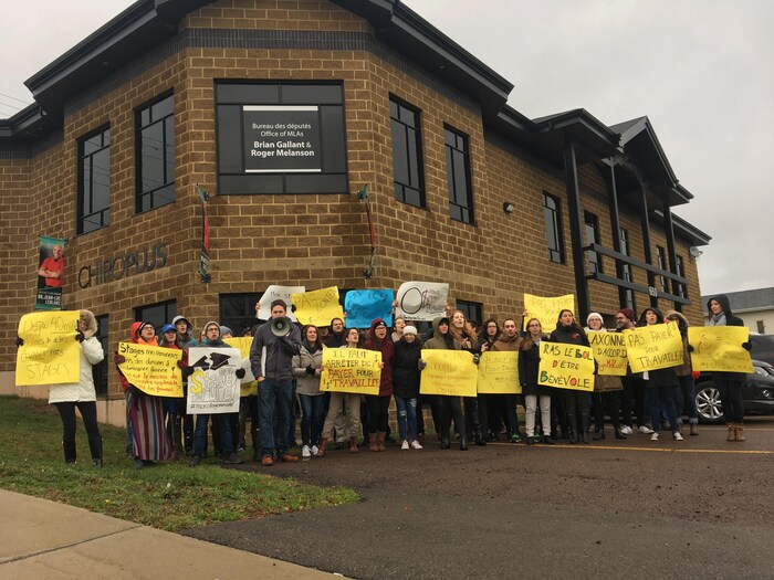 Des étudiants avec des pancartes manifestent devant les bureaux du premier ministre Brian Gallant et du ministre de l'Éducation postsecondaire, Roger Melanson.