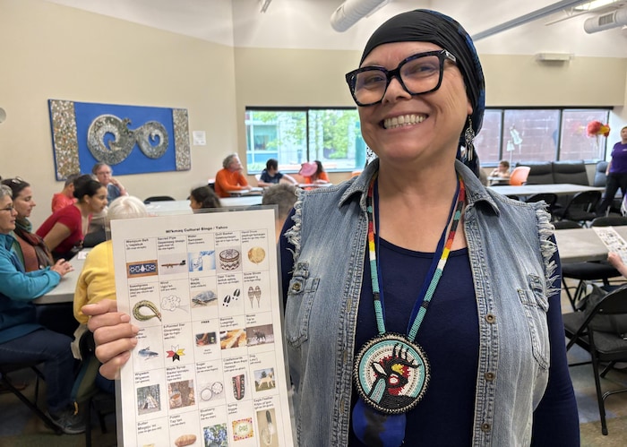 Une femme portant des lunettes à grosse monture noire et un foulard foncé sur la tête fait un grand sourire en montrant une carte sur laquelle il y a plusieurs illustrations.