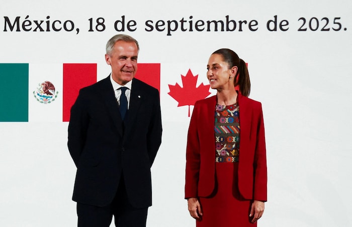 Mark Carney regarde devant lui et Claudia Sheinbaum le regarde. Tous deux sont debout devant un fond blanc décoré des drapeaux du Mexique et du Canada ainsi que du lieu et de la date de leur rencontre, en espagnol.