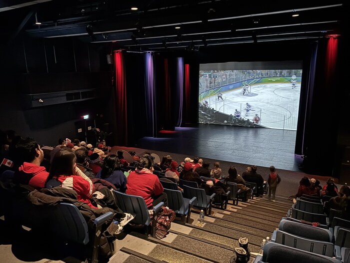 Sur une photo prise du haut de gradins en escalier, on voit des gens assis et un match de hockey qui est diffusé sur un écran géant placé sur la scène d'un théâtre.