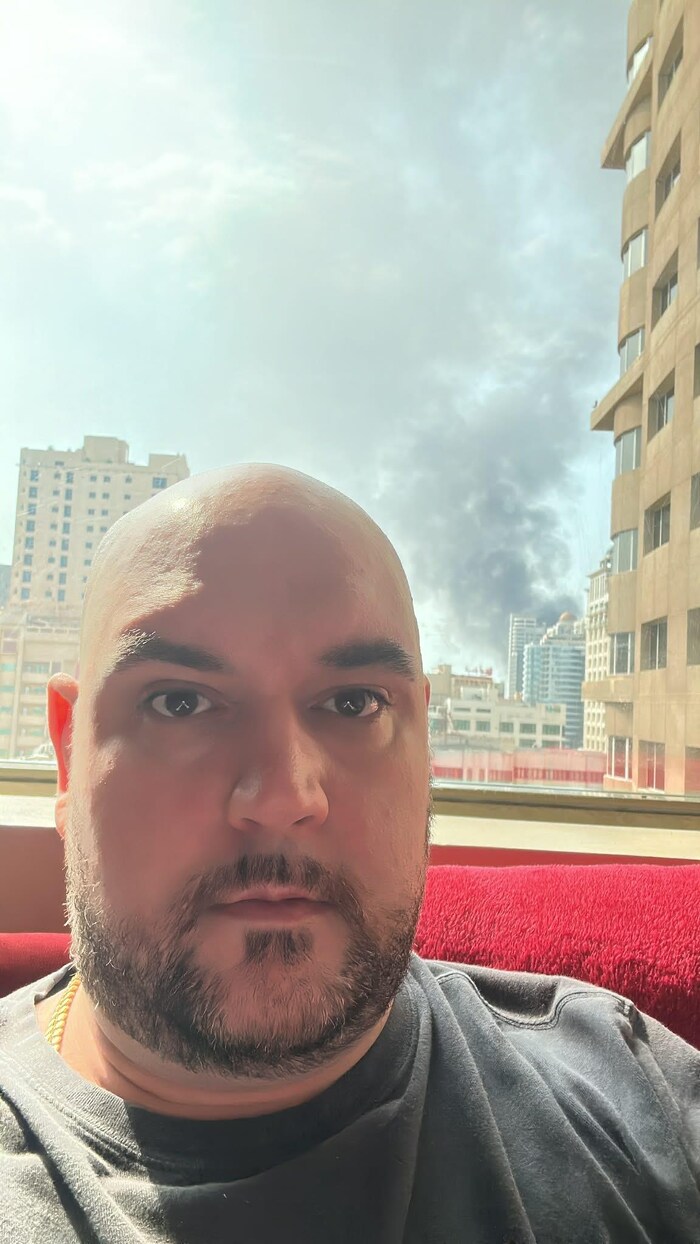 Maxime Proulx fixe l'objectif d'un air sérieux devant une fenêtre à travers laquelle on aperçoit un panache de fumée.