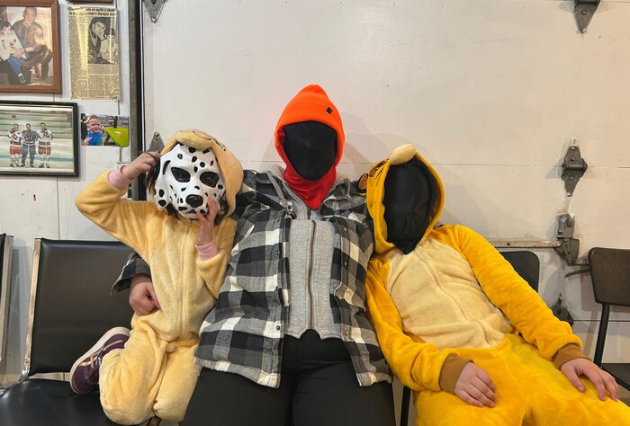 Un adulte et deux enfants sont assis sur des chaises le dos au mur. Deux de ces personnes ont des masques complètement noirs au lieu de leur visage. L'autre personnage est un chien.