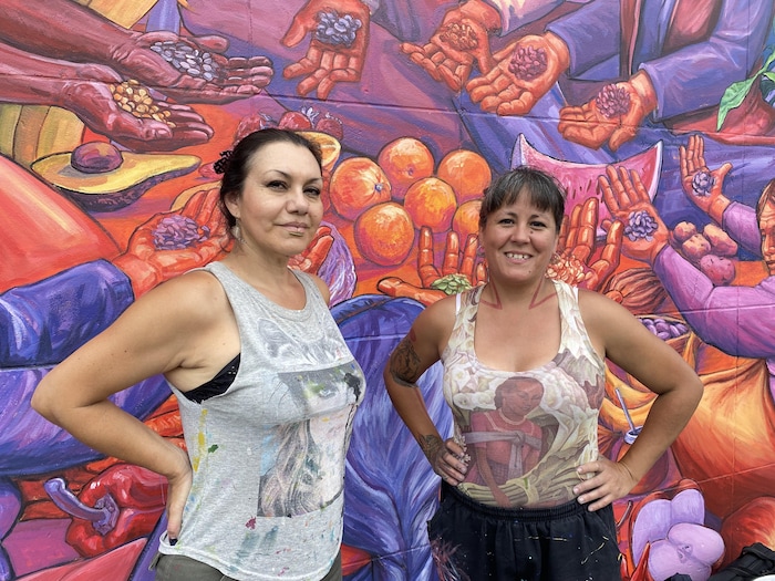 Deux femmes rient ensemble derriére elles, une murale colorée. Il s'agit des artistes Alejandra Zeme (à gauche) et Marila Tarabay, fondatrices du collectif argentin Nereidas R., qui a 20 ans en 2024. Ce sont les muralistes qui ont créé « Guardiennes des sémences » à Montréal. 