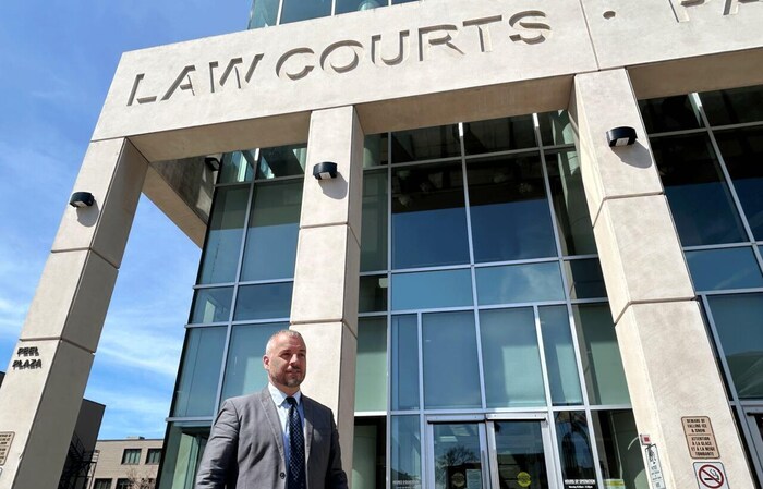 Une photo en contre-plongée d'un homme en complet, debout devant un édifice où les mots «Law Courts - Palais de justice» sont gravés dans la pierre.