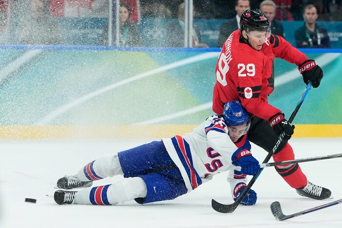 Le joueur des États-Unis tombe à l'horizontale sur son flanc et le joueur canadien est derrière. La photo est obscurcie par la neige qui est déplacée par les patins des hockeyeurs.