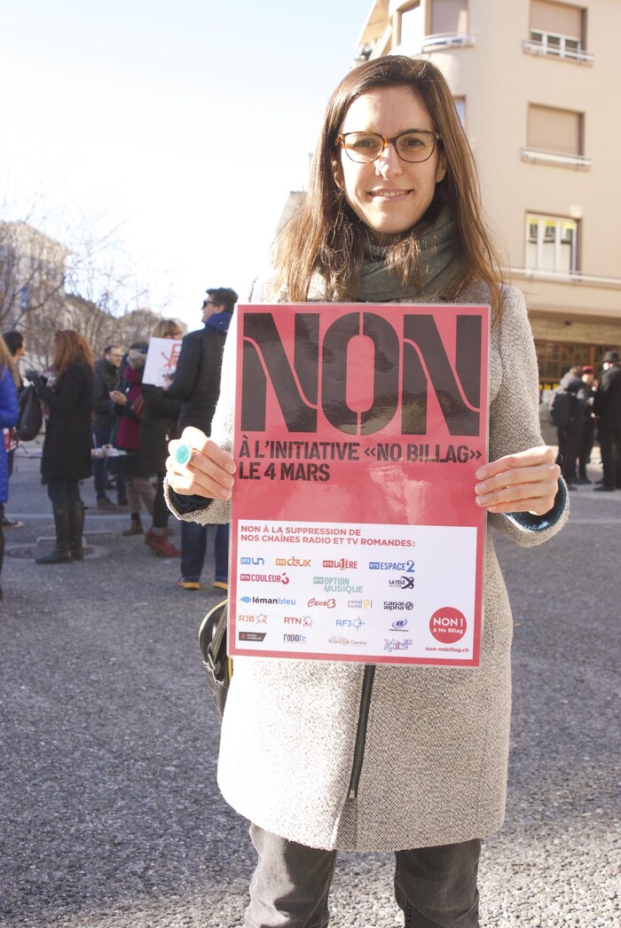 Dans la rue, à un rassemblement populaire, une femme souriante, de face, tient de ses deux mains, au niveau du corps, une affiche sur laquelle on peut lire « Non à l'initiative "No Billag" le 4 mars »