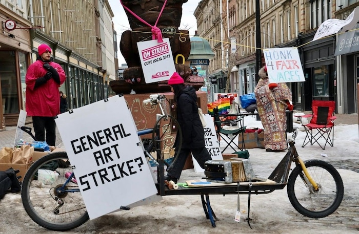 Un engin qui semble être en sculpture en métal à mi-chemin entre un vélo et une trottinette est posé sur une rue piétonnière enneigée et entourée d'affiches indiquant qu'une grève est en cours.