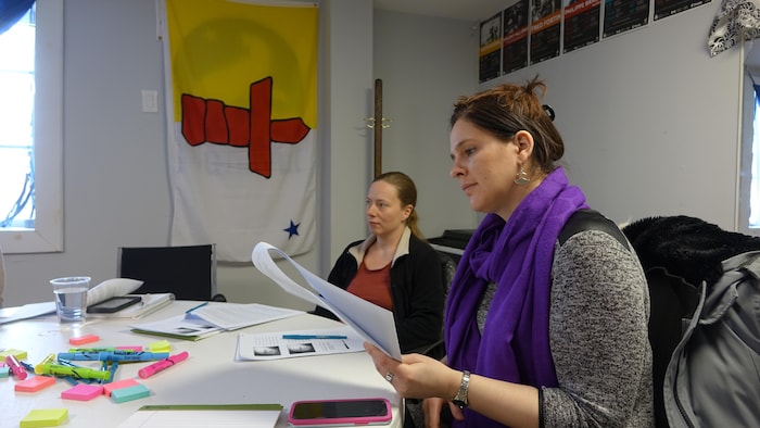 Une femme regarde un document assise à une table avec une autre femme à côté et le drapeau du Nunavut sur le mur.