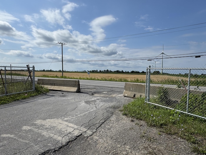 Deux blocs de béton obstruent une entrée clôturée donnant sur une route, des champs et une éolienne.