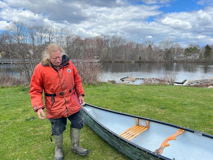 Un homme moustachu aux longs cheveux jaunâtres porte un manteau orange et des bottes en caoutchouc. Il est sur le bord du lac avec une chaloupe.