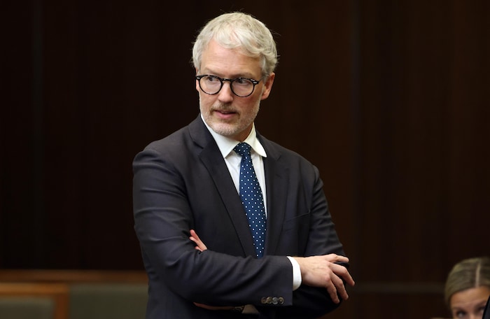 Un homme aux cheveux blancs et aux lunettes à monture ronde est debout, les bras croisés, et jette un regard vers sa droite.