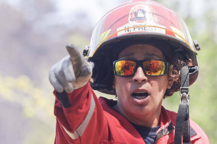 Une pompière casquée, en uniforme, pointe devant elle. Dans ses lunettes fumées, on voit d'autres pompiers devant un feu de forêt.